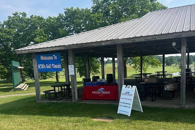 2025 KTA Golf Classic; Outdoor pavilion at a golf course with a "Welcome to KTA's Golf Classic" sign. Tables and a Kentucky Trucking banner are visible, conveying a casual, welcoming tone.