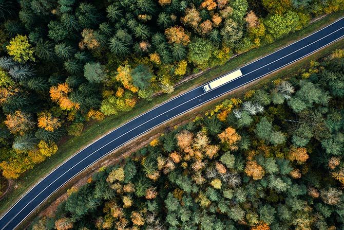 2025 KTA Truck Driving Championship Results; Aerial view of a truck on a winding road through a dense forest with vibrant autumn foliage. The scene conveys a sense of travel and tranquility.