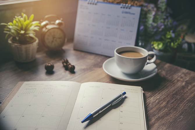 2026 Calendar of Events; A cozy desk scene features a planner open to a monthly view, a pen resting on it, a steaming coffee cup, an old-fashioned alarm clock, and a potted plant.