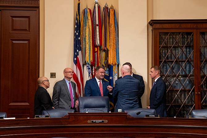 On Capitol Hill, ATA Urges Congress to Strengthen Veterans’ Career Pathways to Trucking; A group of six men in suits converse seriously near a conference table. Behind them, military flags and bookshelves signify an official setting.