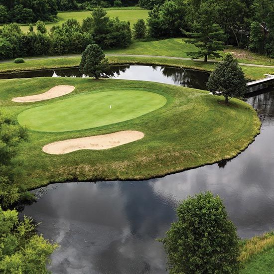 Save the Date! KTA Annual Golf Classic; Aerial view of a golf course with a green surrounded by water, featuring sand bunkers and lush trees. The scene is serene and picturesque.