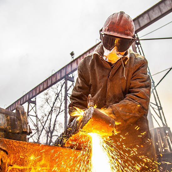 Kentucky Under Construction; A metalworker in protective gear grinds metal, creating bright sparks. The industrial setting is highlighted by steel structures and a cloudy sky.