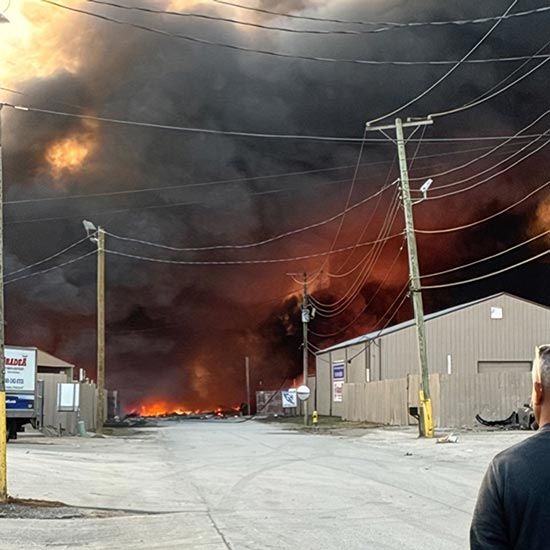The Rebuilding of Redden’s Fleet Maintenance; Massive dark smoke and flames engulf an industrial area, with power lines silhouetted against the sky. A person stands looking at the scene, conveying urgency.