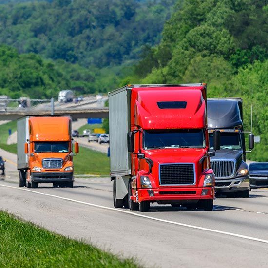 Your Involvement Is What Makes KTA Strong; Three semi-trucks, red, orange, and black, drive on a highway surrounded by lush greenery and hills, evoking a sense of movement and travel.